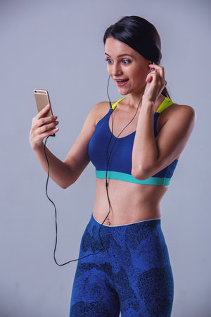 Attractive girl in sportswear is listening to music using a smartphone and smiling, on gray backgroundの写真素材