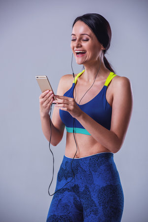 Attractive girl in sportswear is listening to music using a smartphone and smiling, on gray backgroundの写真素材