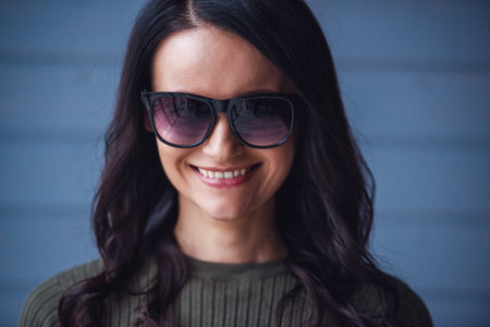Attractive young woman in sun glasses is looking at camera and smiling, on gray backgroundの写真素材