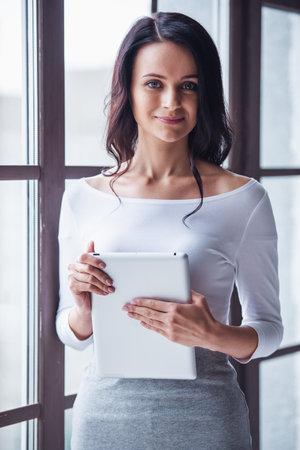 Attractive young woman is holding a digital tablet, looking at camera and smiling while standing near the windowの写真素材