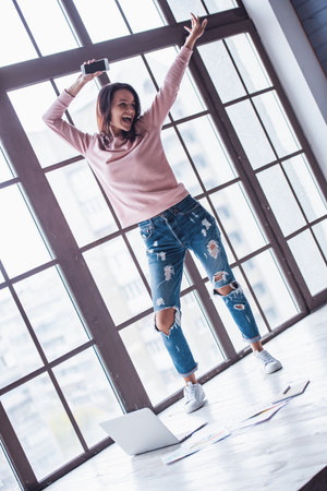 Attractive young woman in casual clothes is dancing and smiling during a break after work with a laptop at homeの写真素材