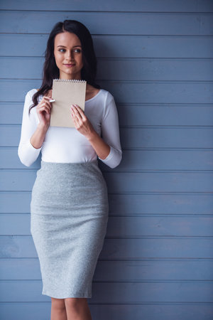 Attractive young woman is holding notepad and pen, thinking and smiling, on gray backgroundの写真素材