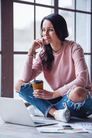 Attractive young woman in casual clothes is drinking coffee, looking away and smiling while working with a laptop at homeの写真素材