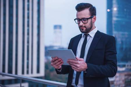 Handsome businessman in suit and glasses is using a digital tablet while standing on the balconyの写真素材