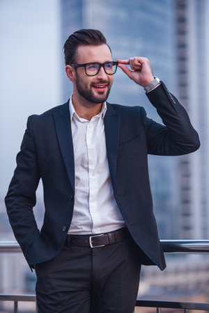 Handsome businessman in suit and glasses is looking away and smiling while standing outdoorsの写真素材