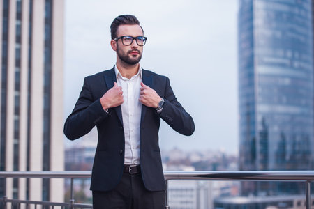 Handsome businessman in suit and glasses is looking away while standing on the balconyの写真素材