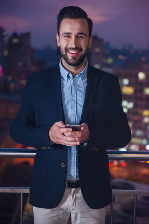 Handsome businessman in suit is holding a smart phone, looking at camera and smiling while standing on the balcony in the eveningの写真素材
