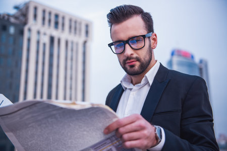 Handsome businessman in suit and glasses is reading newspaper while standing on the balconyの写真素材