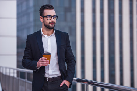 Handsome businessman in suit and glasses is holding a cup of coffee and looking away while standing on the balconyの写真素材