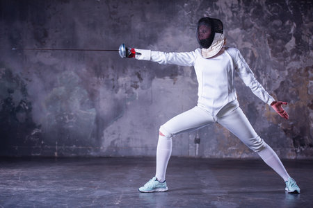Female fencer in protective clothing is using weapon while practicing on dark gray backgroundの写真素材