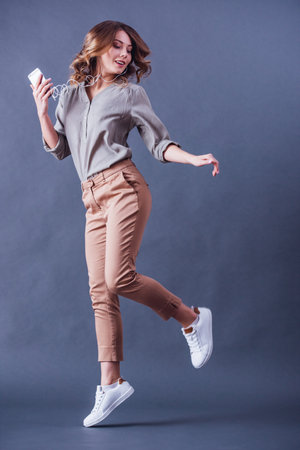 Attractive young woman in earphones is using a smartphone, listening to music, dancing and smiling, on gray backgroundの写真素材