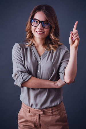 Attractive young woman in smart casual clothes and glasses is pointing upward, looking at camera and smiling, on gray backgroundの写真素材