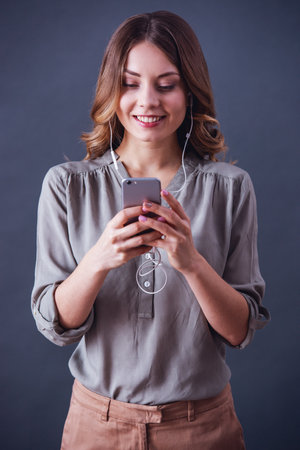 Attractive young woman in smart casual clothes and earphones is using a smartphone and smiling, on gray backgroundの写真素材