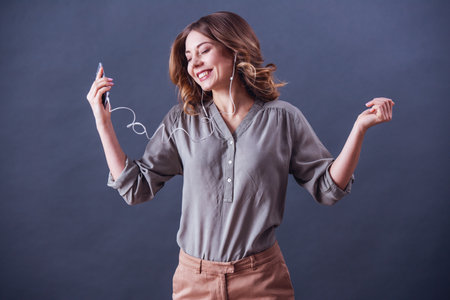 Attractive young woman in smart casual clothes and earphones is using a smartphone, listening to music and smiling, on gray backgroundの写真素材