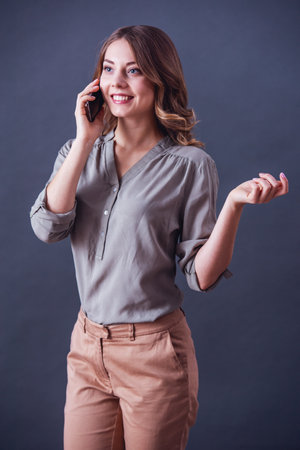 Attractive young woman in smart casual clothes is talking on the mobile phone and smiling, on gray backgroundの写真素材