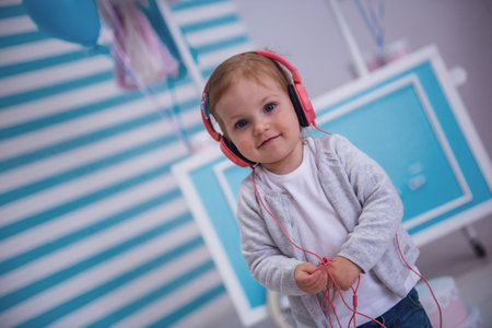 Cute little girl in headphones is looking at camera and smiling while listening to music in the children's roomの写真素材