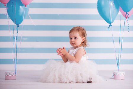 Cute little girl in fluffy white dress is looking away while sitting near balloons, on light backgroundの写真素材