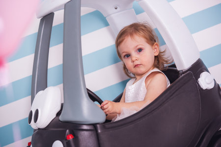 Cute little girl in white dress is looking at camera while sitting in the toy car, on light backgroundの写真素材