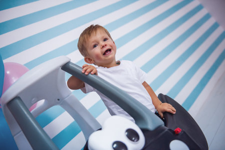 Cute little boy is smiling while sitting in the toy car, on light backgroundの写真素材