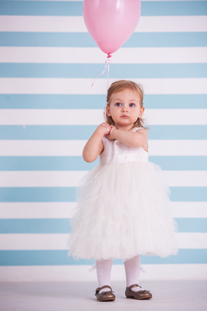 Cute little girl in fluffy white dress is holding a pink balloon and looking away, on light backgroundの写真素材