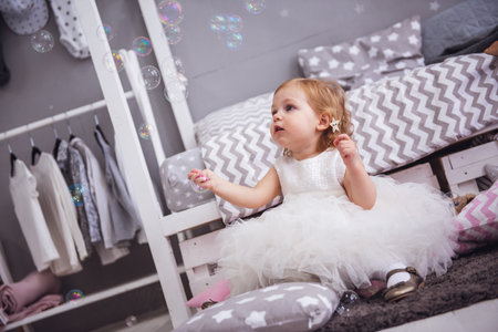 Cute little girl in fluffy white dress is playing with soap bubbles in the children's roomの写真素材
