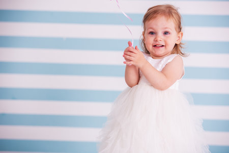 Cute little girl in fluffy white dress is holding a balloon and smiling, on light backgroundの写真素材