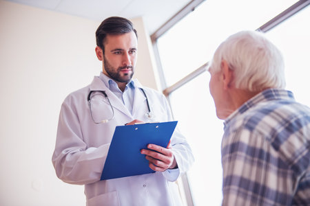 Handsome doctor is talking with an old man and making notes while standing in hospital wardの写真素材