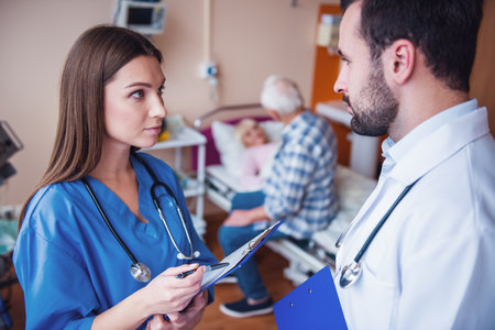 Old man is making a visit to his wife who is lying in bed in the hospital ward, nurse and doctor are talking in the foregroundの写真素材