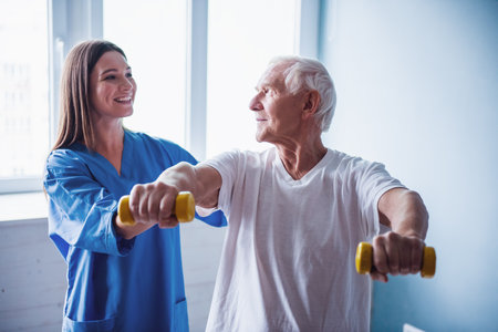 Handsome old man is doing exercises with dumbbells and smiling, in hospital ward. Attractive nurse is helping himの写真素材