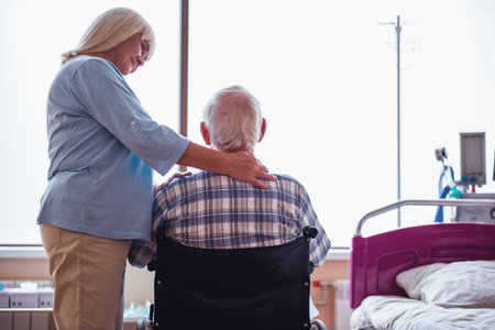 Beautiful old lady is stroking her husband and smiling while he is sitting in the wheelchair in hospital wardの写真素材