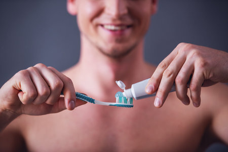 Cropped image of handsome young man applying toothpaste on a toothbrush and smiling, on gray...