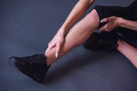Cropped image of girl in sportswear touching her leg feeling pain while sitting on the floor on gray backgroundの写真素材