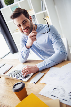 Handsome businessman is holding glasses, looking at camera and smiling while working with the laptop...