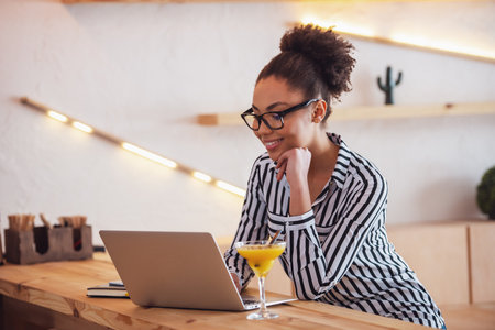 Beautiful Afro American girl in smart casual clothes and glasses is smiling while working with a laptop in cafeの写真素材