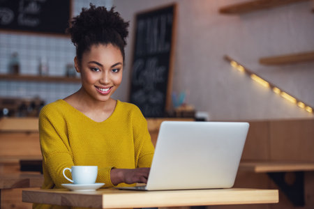 Beautiful Afro American girl in casual clothes is using a laptop, looking at camera and smiling while sitting in cafeの写真素材