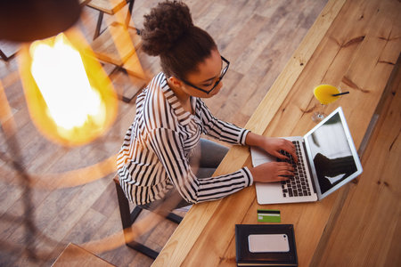 High angle view of beautiful Afro American girl in smart casual clothes and glasses working with a laptop in cafeの写真素材