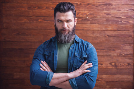 Serious bearded man in casual clothes is standing with crossed arms on a wooden backgroundの写真素材