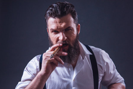 Stylish bearded man in suspenders is smoking a cigar, on dark backgroundの写真素材