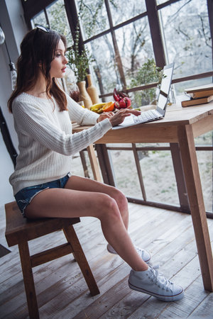 Beautiful young girl in casual clothes is working with a laptop while sitting at the wooden table at homeの写真素材
