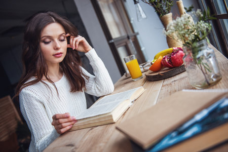 Beautiful young girl in casual clothes is reading a book while sitting at the wooden table at homeの写真素材
