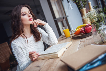 Beautiful young girl in casual clothes is reading a book and looking out the window while sitting at the wooden table at homeの写真素材