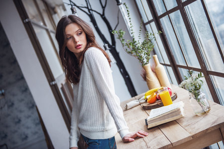 Beautiful young girl in casual clothes is looking away while leaning on wooden table at homeの写真素材