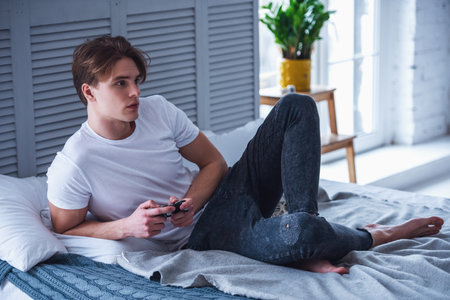 Handsome young man in casual clothes is playing video game console while sitting on his bed at homeの写真素材