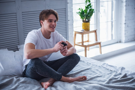 Handsome young man in casual clothes is playing video game console and smiling while sitting on his bed at homeの写真素材