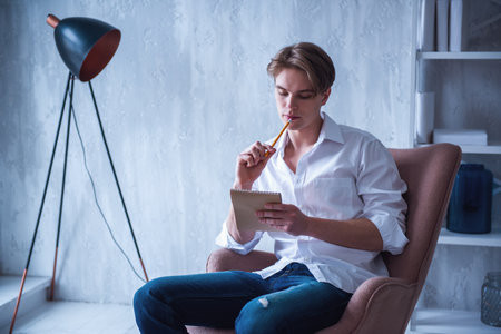 Handsome young man in jeans and white shirt is thinking and making notes while sitting in armchair at homeの写真素材