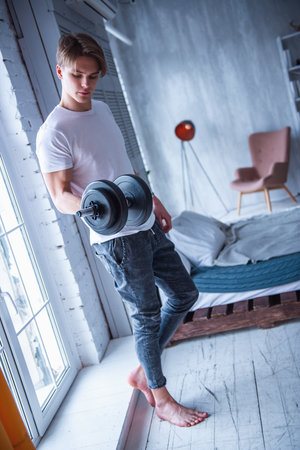 Handsome young man in casual clothes is working out with a dumbbell in his room at home, full-lengthの写真素材