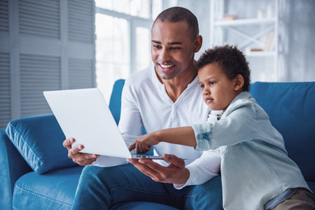 Happy father and son in casual clothes are using a laptop while sitting on couch at home, dad is smilingの写真素材