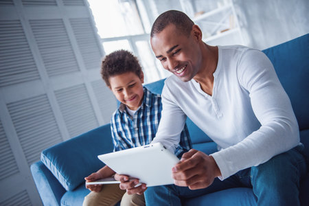 Happy father and son in casual clothes are using digital tablets and smiling while sitting on couch at homeの写真素材
