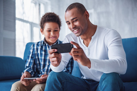 Happy father and son in casual clothes are using gadgets and smiling while sitting on couch at homeの写真素材