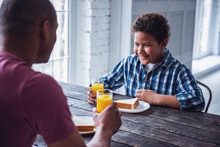 Handsome father and son in casual clothes are eating at the wooden table together at home, boy is smilingの写真素材